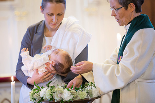 Kuvassa ristiäiset. Pappi kastaa kummin sylissä olevaa vauvaa.
